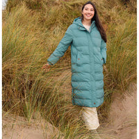 A woman wearing a long teal puffer coat and cream skirt stands on sandy dunes surrounded by tall grass.