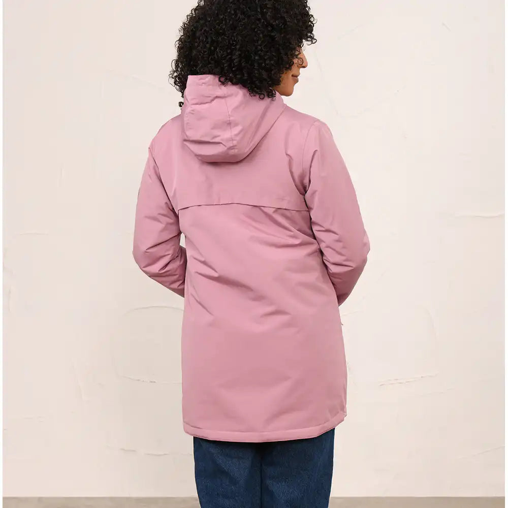 A woman viewed from the back wearing a dusty pink hooded jacket and blue jeans, standing against a plain off-white wall.