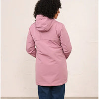 A woman viewed from the back wearing a dusty pink hooded jacket and blue jeans, standing against a plain off-white wall.