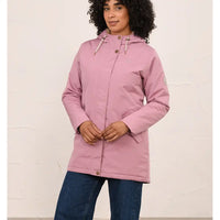 A woman wearing a dusty pink hooded raincoat with a high collar and front zipper, paired with blue jeans, standing against a textured off-white wall.