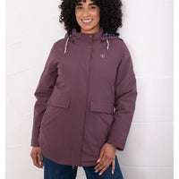 A woman wearing a dusty plum hooded jacket with front flap pockets and drawstrings, paired with blue jeans, standing against a white brick wall.