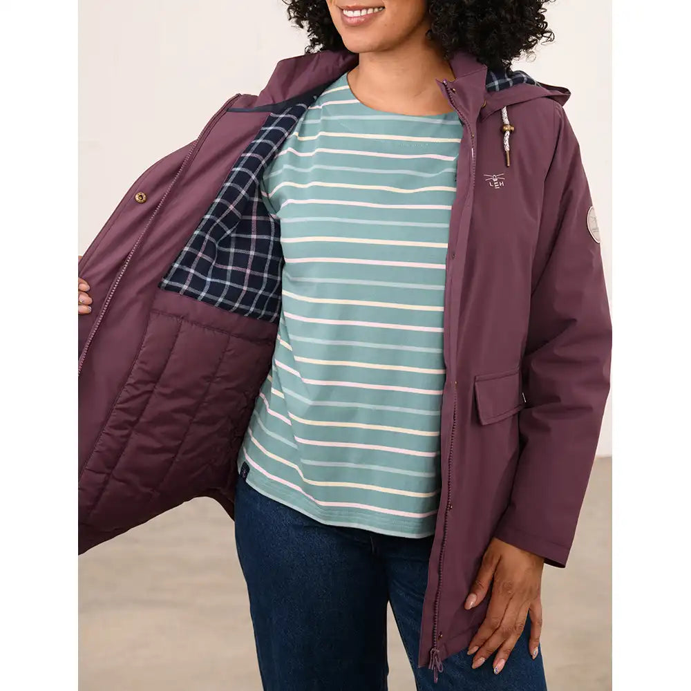 A woman wearing a plum purple hooded puffer jacket with a green and white striped shirt underneath, holding the jacket open to show the plaid lining. The jacket has a small embroidered logo on the left chest.