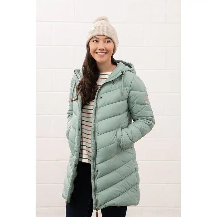 Woman wearing a light green puffer coat and white beanie against a white brick wall.