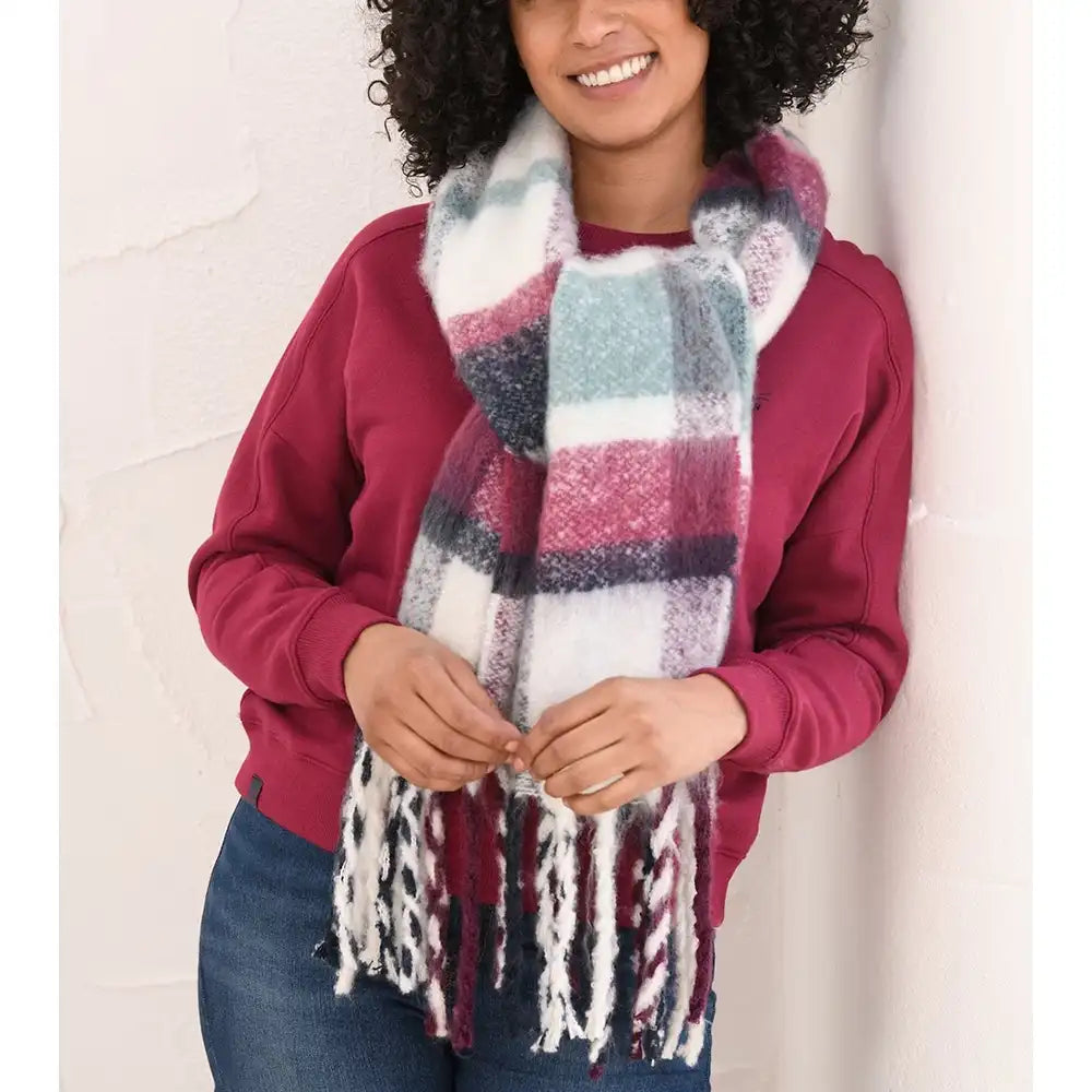 A woman wearing a chunky, fuzzy plaid scarf in white, burgundy, and teal, styled with a burgundy sweatshirt and blue jeans against a white textured wall.