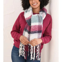A woman wearing a chunky, fuzzy plaid scarf in white, burgundy, and teal, styled with a burgundy sweatshirt and blue jeans against a white textured wall.
