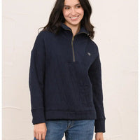 A woman wearing a navy blue quilted quarter-zip pullover sweater with a small embroidered logo on the chest, paired with blue jeans, standing against a textured off-white wall.