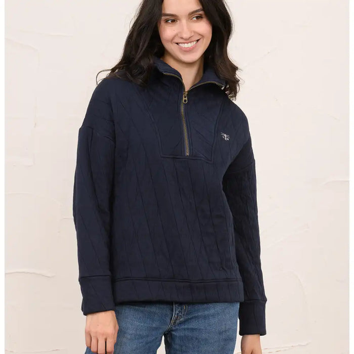 A woman wearing a navy blue quilted quarter-zip pullover sweater with a small embroidered logo on the chest, paired with blue jeans, standing against a textured off-white wall.