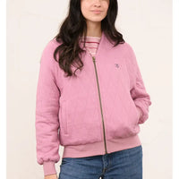 A woman wearing a pink quilted zip-up bomber jacket with a small embroidered logo on the chest, paired with a striped shirt and blue jeans, against a light beige background.