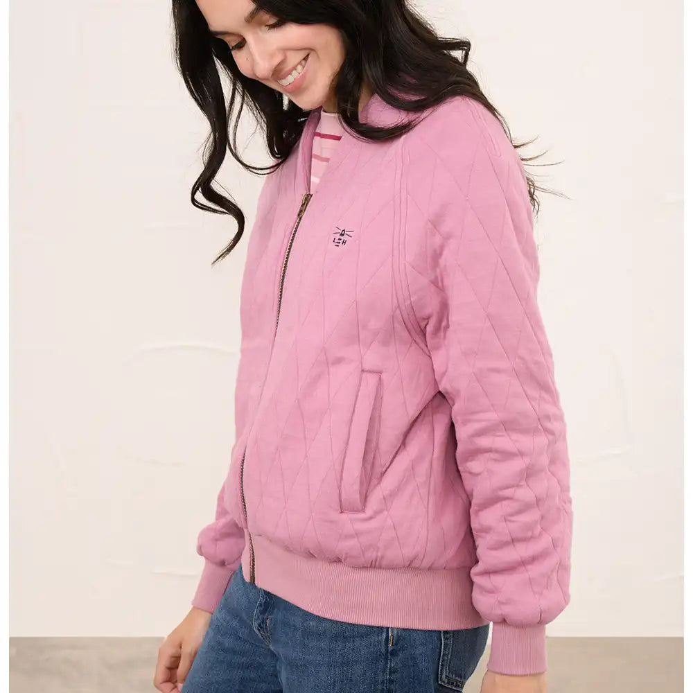A woman wearing a pink quilted zip-up bomber jacket with a small embroidered logo on the chest, paired with blue jeans, against a plain off-white wall.