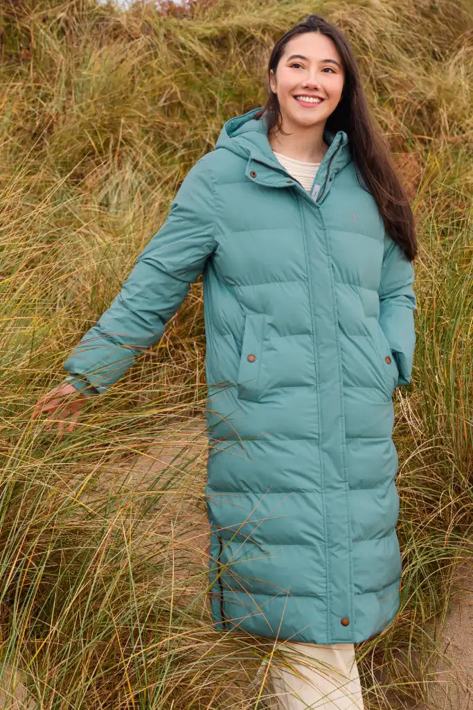 A woman wearing a long teal-green hooded puffer coat stands outdoors in tall dry grass, smiling and posing with one arm extended.
