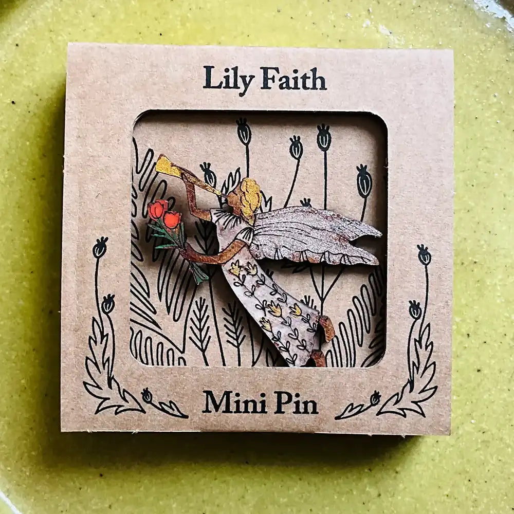 A mini pin by Lily Faith shown in its kraft paper packaging, featuring a wooden pin of a fairy with wings and a leafy branch.