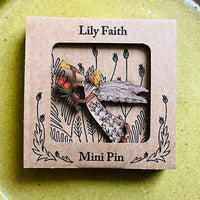 A mini pin by Lily Faith shown in its kraft paper packaging, featuring a wooden pin of a fairy with wings and a leafy branch.