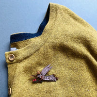 A close-up of a mustard yellow knit cardigan sweater with a dark blue ribbed collar, wooden buttons, and a small embroidered fairy pin on the chest, laid flat on a blue-gray background.