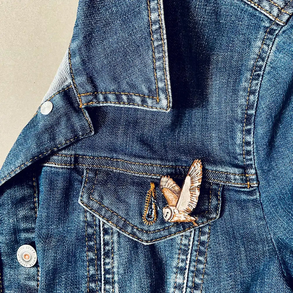 A close-up of a blue denim jacket showing a front pocket with a small barn owl pin attached. 