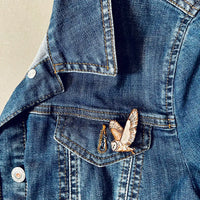 A close-up of a blue denim jacket showing a front pocket with a small barn owl pin attached. 