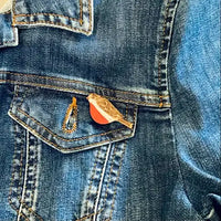A close-up of a blue denim jacket showing a front pocket with a small robin pin attached. 