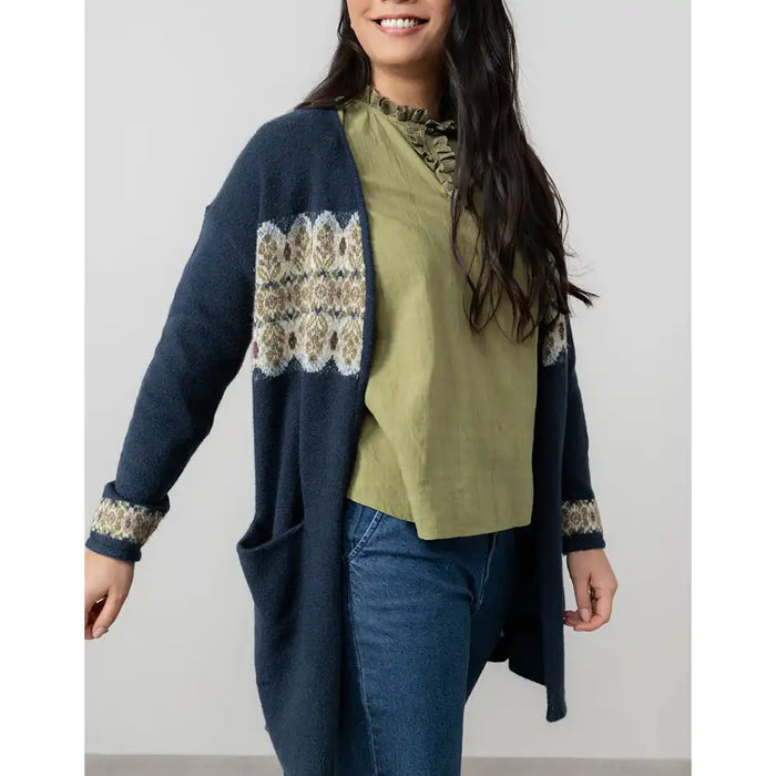 Woman wearing a navy cardigan with patterned pockets, green top, and blue jeans against a white background
