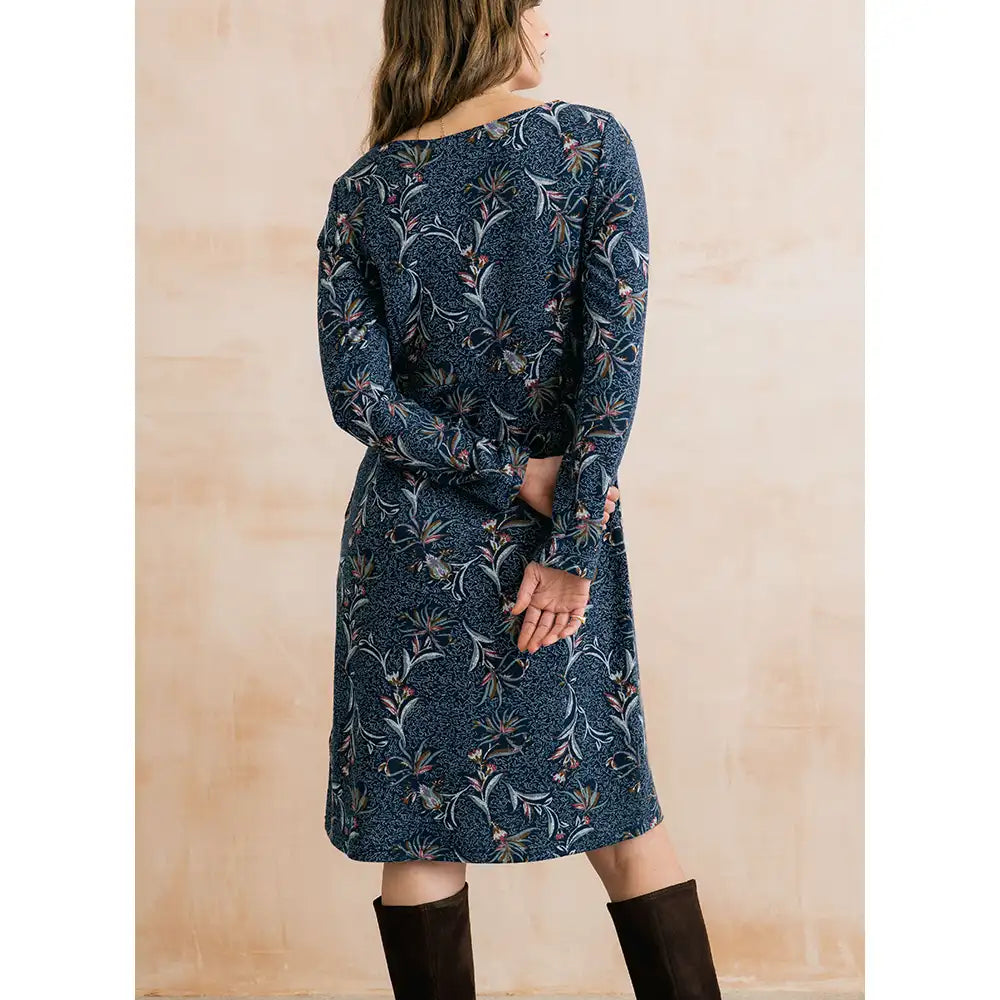 A woman wearing a long-sleeve knee-length dress with a dark blue and black floral pattern, shown from the back against a warm beige background.