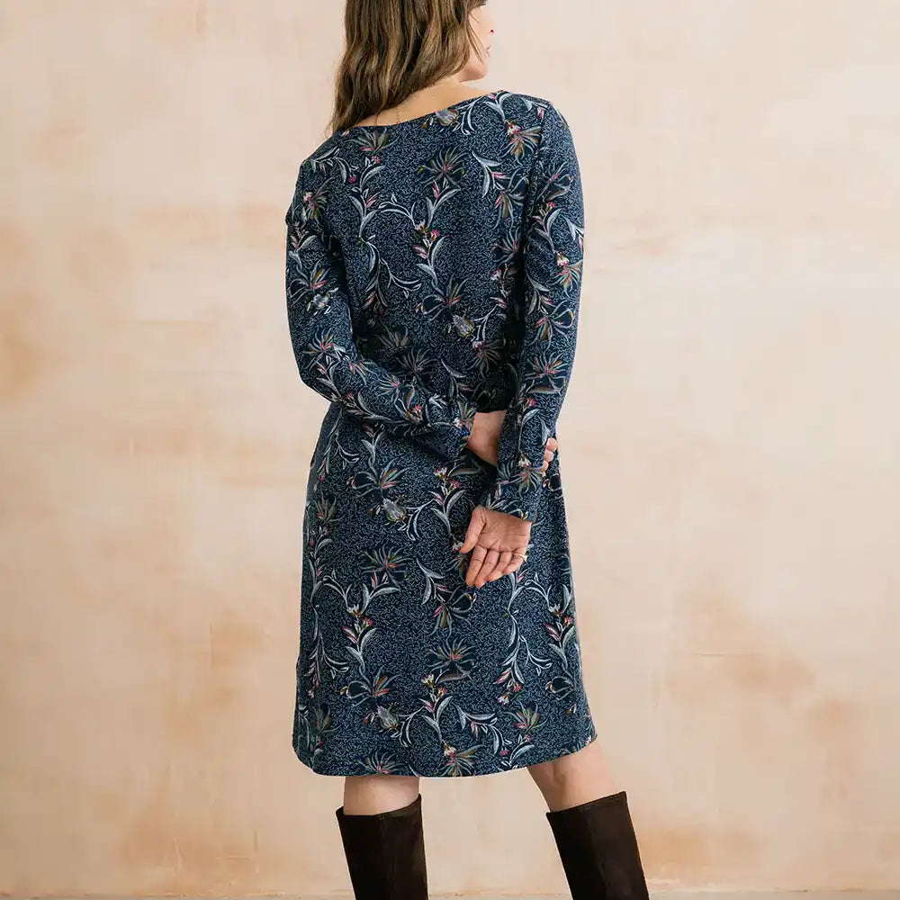 A woman wearing a long-sleeve navy blue dress with a detailed botanical pattern, shown from the back against a textured beige wall.
