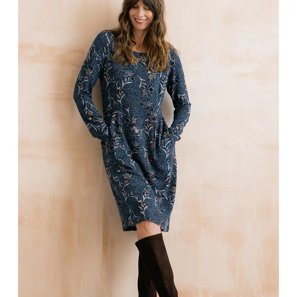 A woman wearing a long-sleeve knee-length blue dress with a white floral pattern and black over-the-knee boots, standing against a warm beige wall.