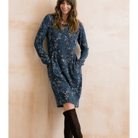 A woman wearing a long-sleeve knee-length blue dress with a white floral pattern and black over-the-knee boots, standing against a warm beige wall.