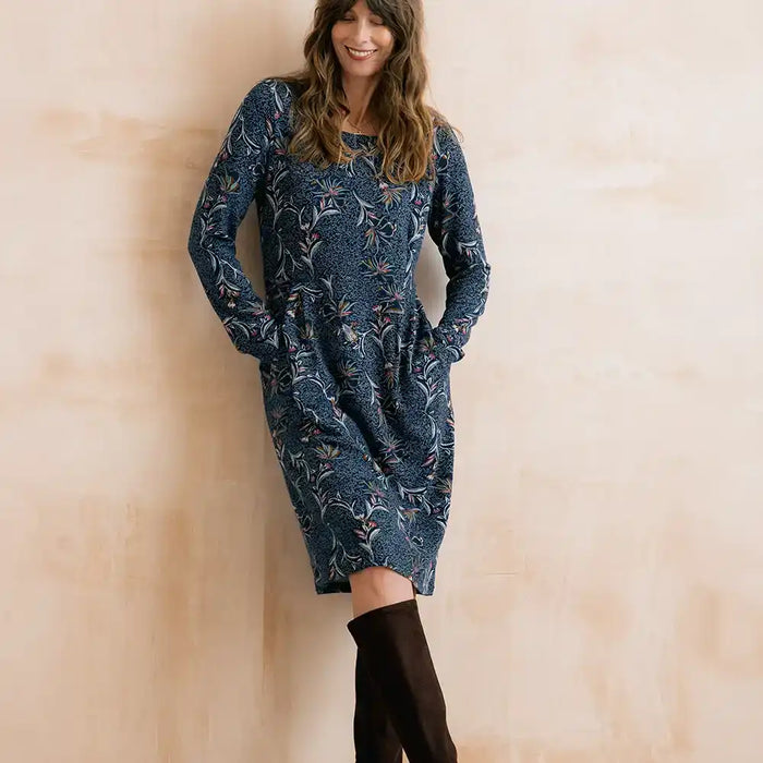 A woman wearing a long-sleeve knee-length blue dress with a white floral pattern and black over-the-knee boots, standing against a warm beige textured wall.