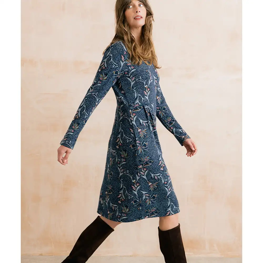 A woman wearing a long-sleeve knee-length dress with a blue floral and botanical pattern, paired with black knee-high boots, posing against a textured beige wall.