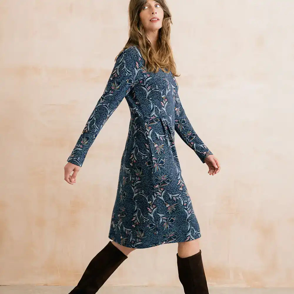 A woman wearing a long-sleeve knee-length blue dress with a white floral pattern and black over-the-knee boots, standing against a textured beige wall.