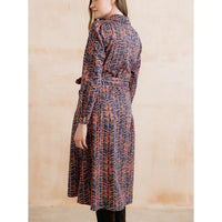 A woman wearing a long-sleeved belted wrap dress with a navy blue and rust-orange abstract leaf pattern, shown from a side-back angle against a textured beige wall.