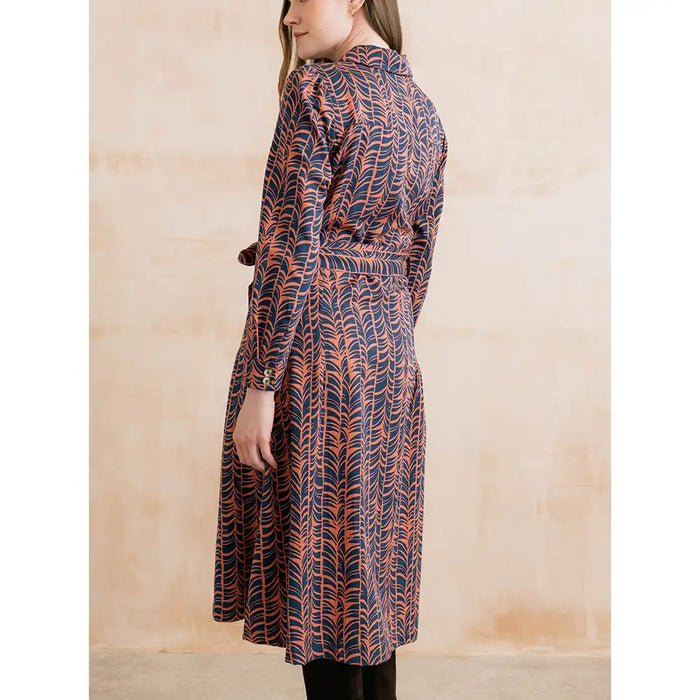 A woman wearing a long-sleeved belted wrap dress with a navy blue and rust-orange abstract leaf pattern, shown from a side-back angle against a textured beige wall.