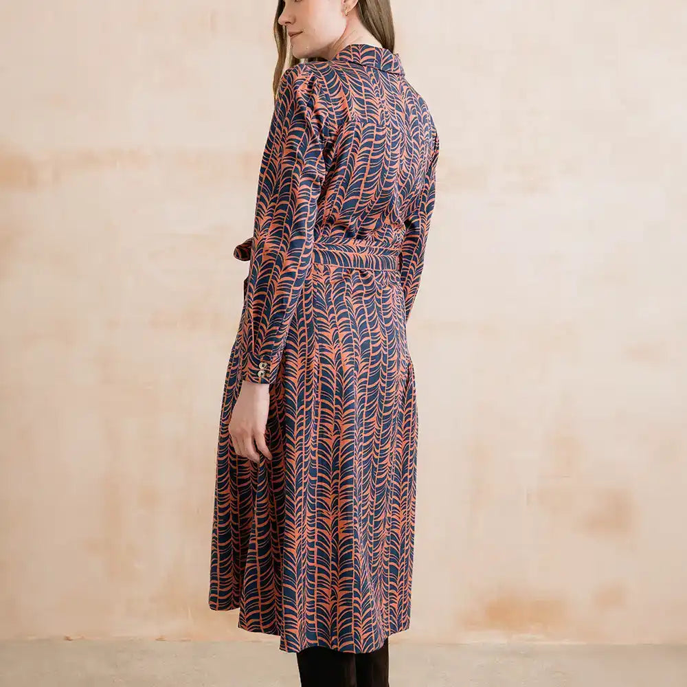 A woman wearing a long-sleeve belted shirt dress with a navy blue and orange abstract leaf pattern, shown from a side-back angle against a textured beige wall.