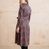 A woman wearing a long-sleeve belted shirt dress with a navy blue and orange abstract leaf pattern, shown from a side-back angle against a textured beige wall.