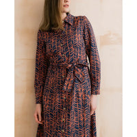 A woman wearing a long-sleeve button-front dress with a self-tie waist belt, featuring an abstract wavy pattern in navy blue and rust orange, set against a warm beige background.