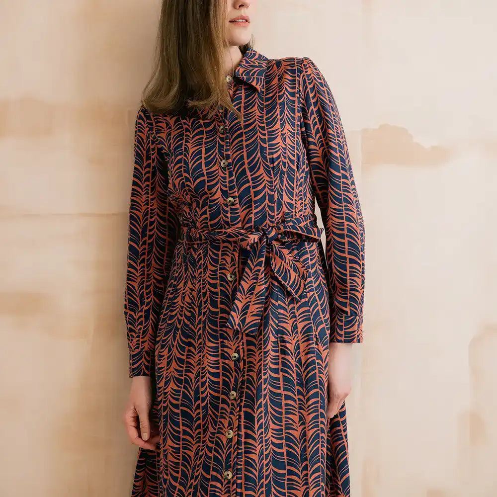 A woman wearing a long-sleeve button-front dress with a self-tie waist belt, featuring an abstract wavy pattern in navy blue and rust orange, posed against a textured beige wall.