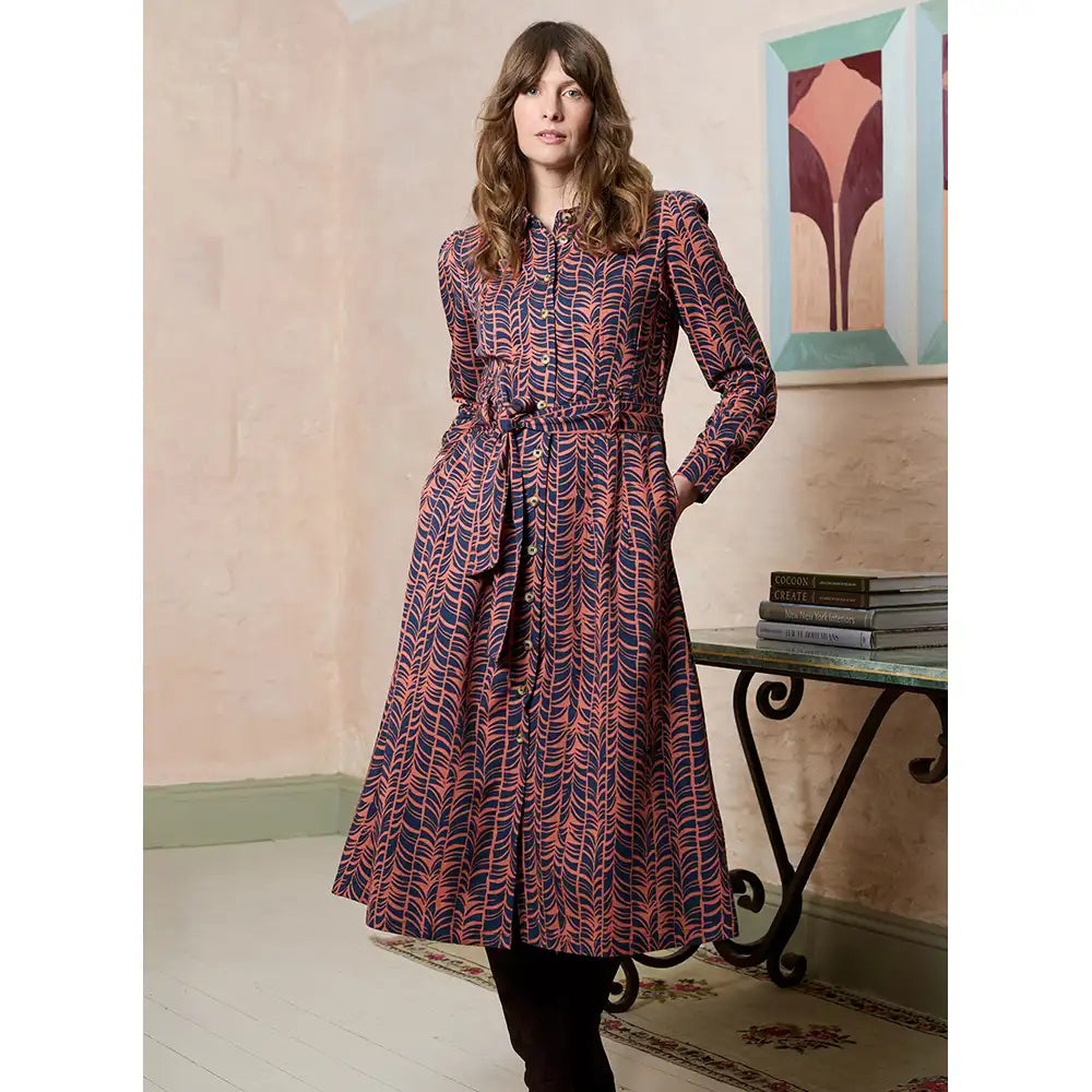 A woman models a long-sleeved midi dress with a red and navy blue abstract wavy pattern, featuring a tie waist belt. She is standing in an indoor setting with a table and framed art in the background.