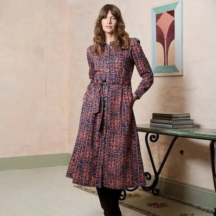 A woman models a long-sleeved midi dress with a red and navy blue abstract wavy pattern, standing in a room with a console table and framed art.