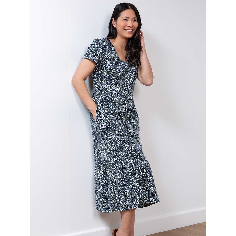 Dark hair model with right hand in pocket at waist, left hand in hair wearing a navy blue dress that is mid-calf length with short sleeves and v neckline 