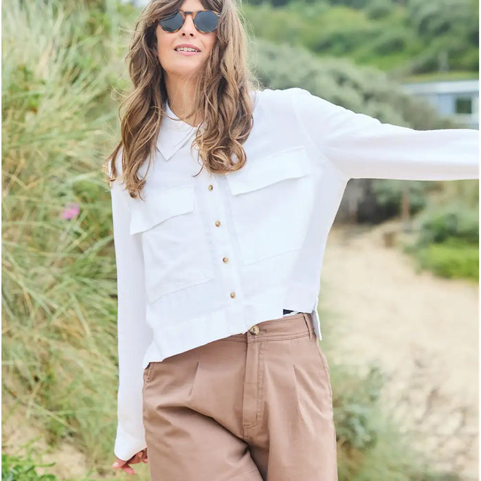 Woman wearing a white blouse and brown pants outdoors with greenery in the background
