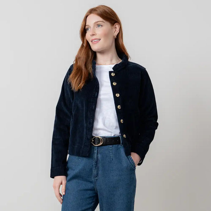 Woman wearing a black jacket with gold buttons, white shirt, and blue jeans on a light gray background