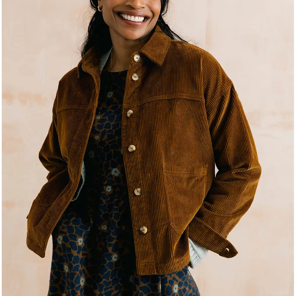 A smiling woman wears a brown corduroy button-front jacket over a dark patterned dress, photographed against a soft beige wall.