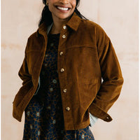 A smiling woman wears a brown corduroy button-front jacket over a dark patterned dress, photographed against a soft beige wall.