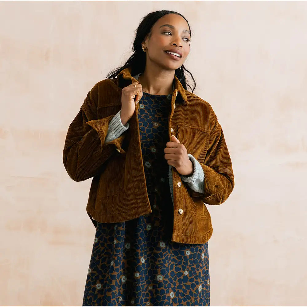 A woman wearing a brown corduroy jacket over a blue patterned dress, posing against a light beige textured background.