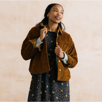 A woman wearing a brown corduroy jacket over a blue patterned dress, posing against a light beige textured background.