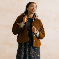 A woman wearing a brown corduroy jacket over a blue patterned dress, standing against a warm beige textured background.