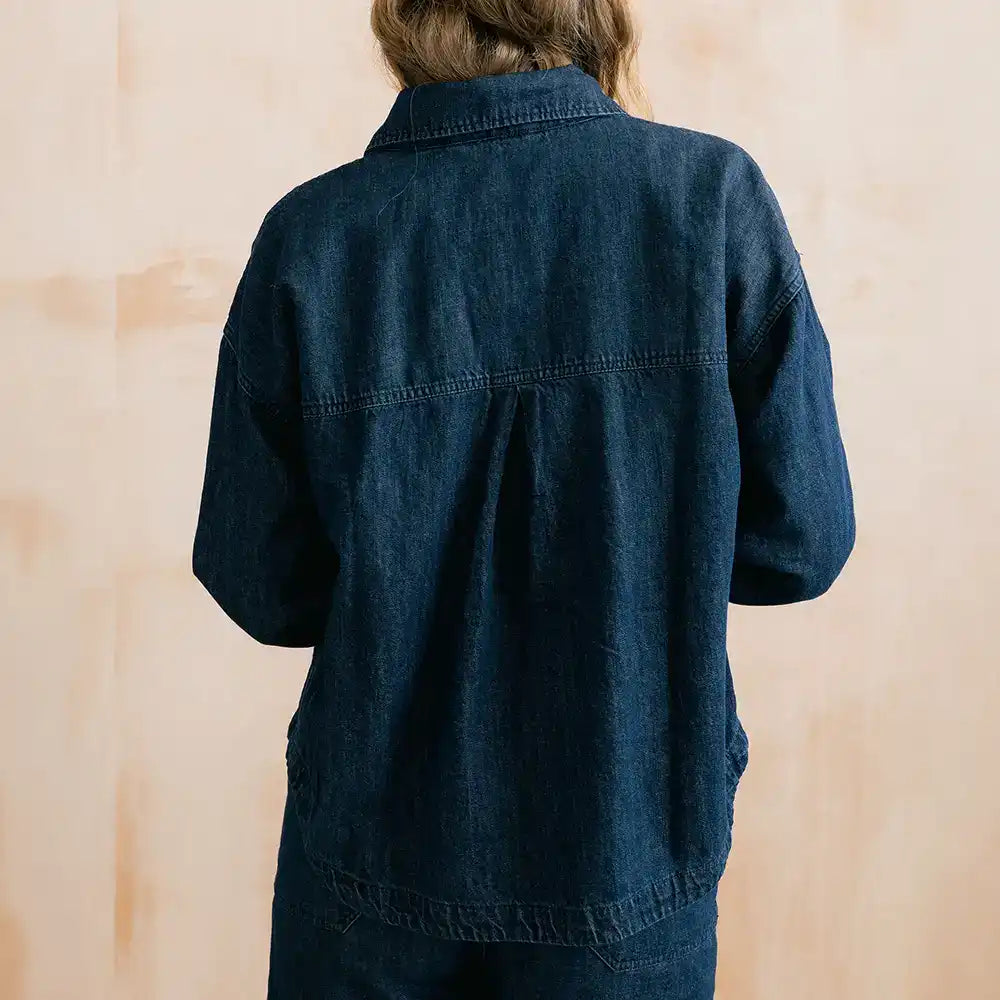 A person shown from the back wearing a dark indigo denim jacket or overshirt against a light beige textured wall.