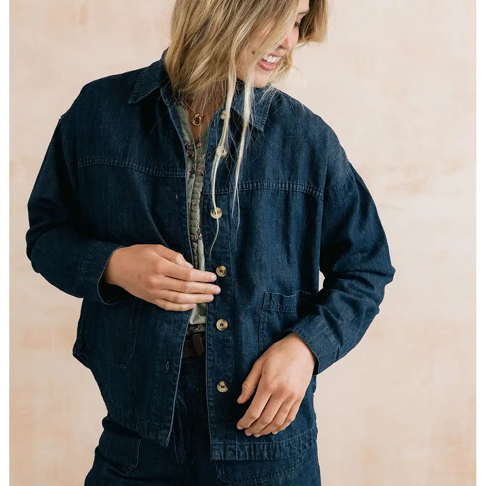 A woman wearing an oversized dark indigo denim button-up shirt jacket over a patterned shirt, standing against a soft beige textured background.