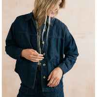 A woman wearing an oversized dark indigo denim button-up shirt jacket over a patterned shirt, standing against a soft beige textured background.