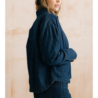 A woman wearing an oversized dark indigo denim shirt jacket, shown from a side profile against a warm beige textured background.