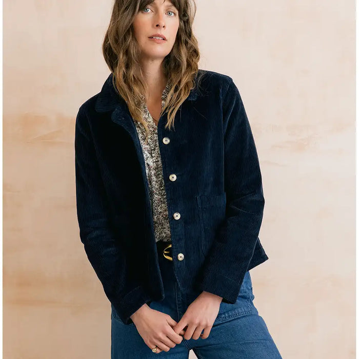 A woman wearing a dark navy corduroy jacket over a patterned top and blue jeans, standing against a warm beige textured wall.