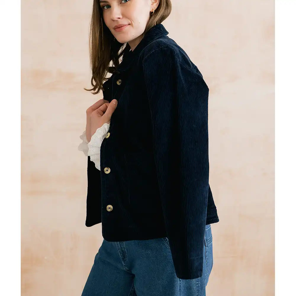 A woman wearing a dark navy corduroy jacket with gold buttons over a white ruffled shirt and blue jeans, standing against a light beige textured wall.